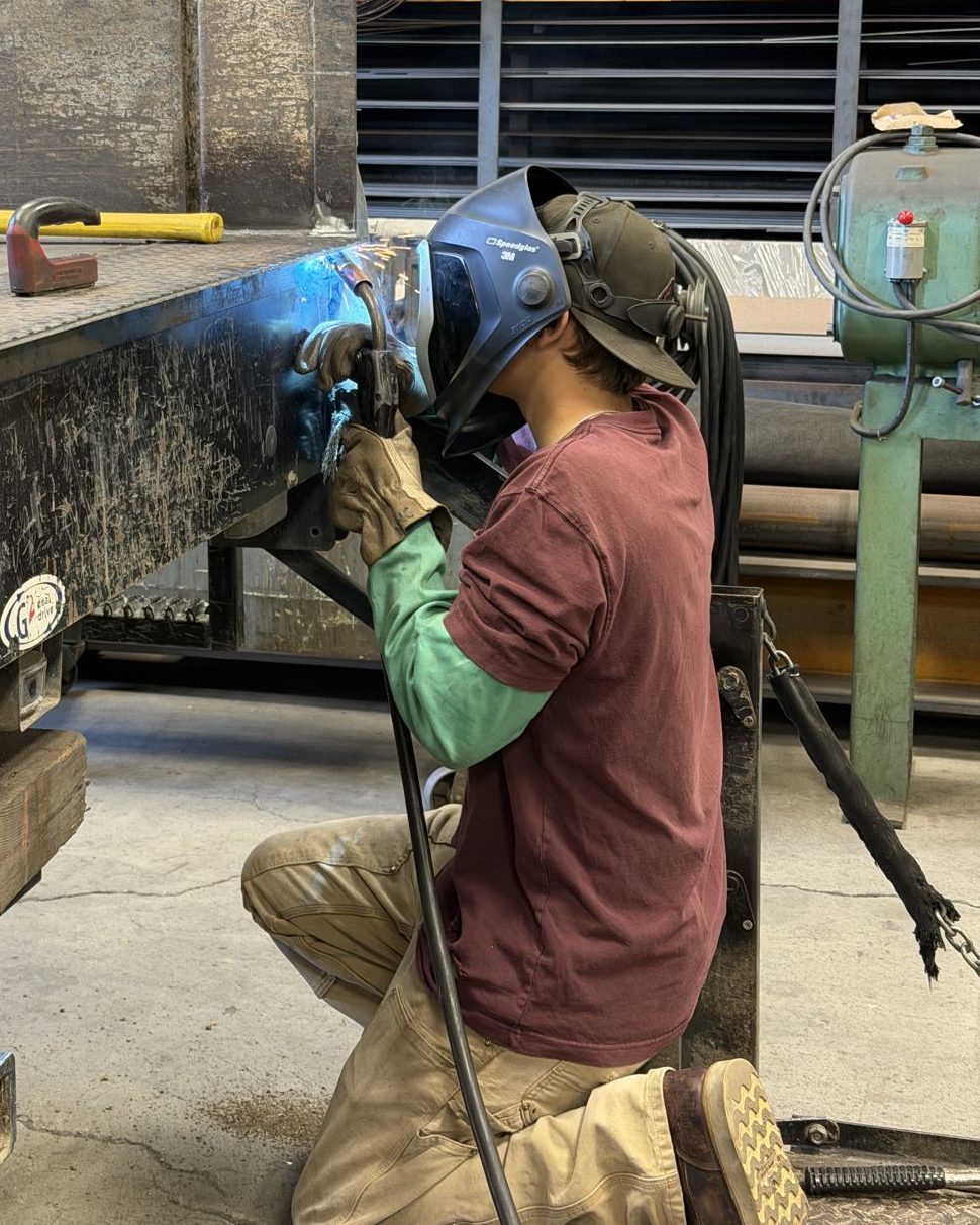 Welder in a helmet kneeling to weld a repair on a flatbed truck