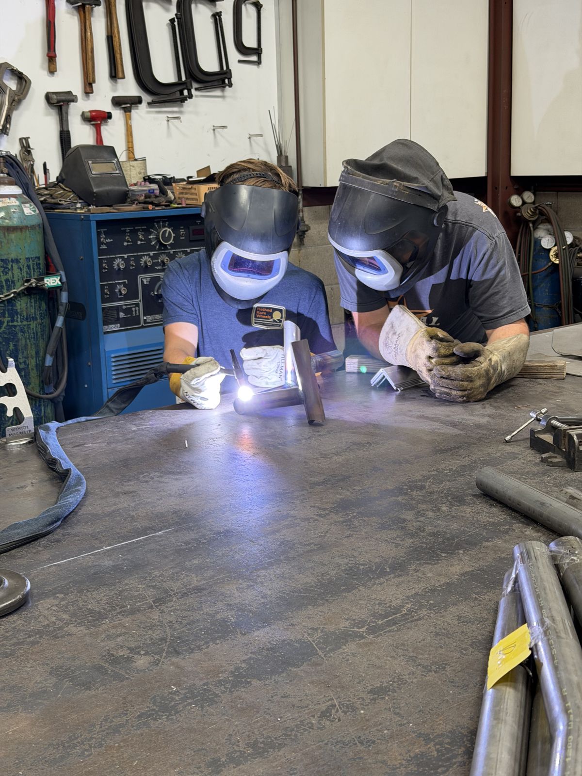 Two welders in helmets welding a steel part at the shop bench